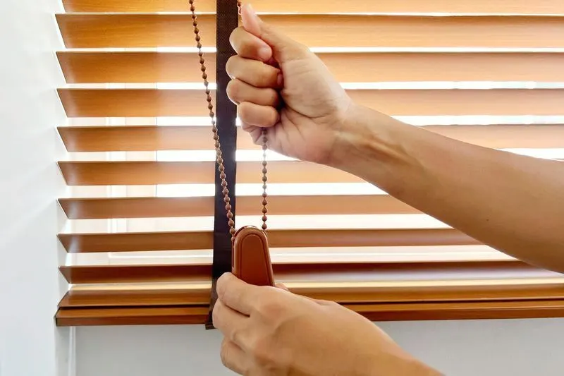 Hand adjusting wood blinds on a sunny window